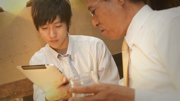 居酒屋でタブレットを操作する若いサラリーマンと、グラスを持ちながら説明を聞く上司