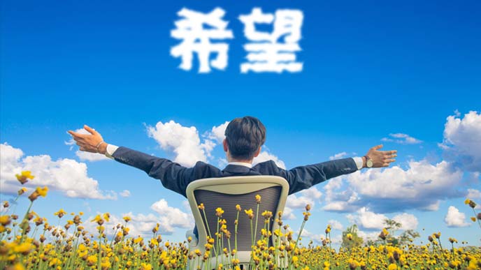草原でオフィスチェに座りながら両手を広げる男性の後ろ姿